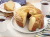 Fotografia em tons de azul e amarelo em uma bancada de madeira com um pano branco com listras azuis e amarelas. Ao centro, um prato branco redondo com o bolo branco em cima dele. Ao lado, xícaras de café e um prato com uma fatia do bolo.