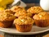 A imagem mostra vários muffins de banana com uma cobertura crocante, dispostos em um prato claro sobre uma mesa rústica de madeira. Os muffins têm uma cor dourada e textura levemente crocante por fora, com aparência macia por dentro.