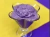 sobre um fundo azul, amarelo e branco, dentro uma taça de vidro transparente está um sorvete cremoso em tons de roxo com pedacinhos de uva.