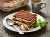 Em cima de uma mesa, prato com porção de tiramisu feito com biscoitos champanhe, creme branco cremoso, polvilhado com cacau em pó.