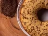 Fotografia vista de cima de um bolo formigueiro inteiro sobre um prato branco que está apoiado em uma mesa de madeira. Ao lado do prato tem um pouco de granulado de chocolate.