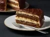Fotografia de uma fatia de bolo de chocolate com camadas, coberta por ganache brilhante e cremoso, servida em um prato branco. O bolo apresenta duas camadas de um bolo de chocolate fofinho e um recheio cremoso.