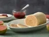 Imagem de um prato oval com o bolo de rolo e duas fatias cortadas. Ao fundo, uma pote de goiabada e goiabas ao lado.
