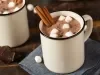 Imagem de uma caneca branca com chocolate quente, pedaços de marshmallow e 2 pedaços de canela em pau.