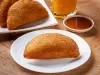 Fotografia de um risole de presunto e queijo em um prato raso branco sobre a mesa de madeira. Ao fundo tem suco de laranja, o molho e mais risoles.