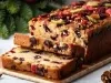 Fotografia de um bolo de frutas festivo, exibido em uma tábua de madeira. O bolo, denso e recheado com frutas secas e nozes, é cortado para mostrar seu interior colorido. O fundo sugere um tema natalino, com ramos de pinheiro.