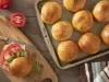 Imagem vista de cima de uma forma com vários pães redondos de batata-doce. Do lado esquerdo, um lanche montado com o pão, presunto, queijo, tomate e rúcula.