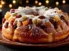 Fotografia de um bolo de frutas em formato de anel, adornado com frutas cristalizadas e polvilhado com açúcar de confeiteiro. O fundo desfocado com luzes quentes evoca um clima natalino e festivo.