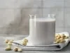 Fotografia em tons claros de um copo de vidro com uma bebida vegetal de castanha de caju apoiado em um pano branco dobrado com algumas listras escuras, que está sobre uma mesa de cimento queimado. Por cima da mesa e do pano tem castanhas espalhadas.