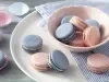 Imagem de macarons rosa e azul em um pote rosa claro sobre um prato branco. Ao lado mais macarons em cima do prato.