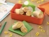 Fotografia de um recipiente laranja quadrado e fundo com enroladinhos de pão com cenoura e ovo. Dentro do recipiente tem um papel verde e branco, e ao lado tem mais dois rolinhos sobre um papel verde dobrado e uma mesa de madeira.