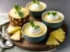 Fotografia de tigelas azuis, com creme amarelo, cobertura de chantilly e folhas de hortelã. Um dos doces é servido em uma metade de abacaxi, rodeado por fatias de abacaxi fresco em uma tábua de madeira.