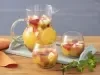 Fotografia em tons de laranja em uma bancada de madeira, um paninho laranja, uma tábua azul de madeira, uma jarra de vidro com frutas e espumante e ao lado dois copos de vidro com a bebida servida dentro deles. E folhas de hortelã para decorar a mesa.