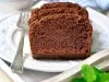 Fatias de bolo de chocolate fofinho em um prato decorado com flores azuis. Um garfo prateado ao lado e uma folha de hortelã adicionam charme. O fundo desfocado com um pano xadrez cria uma atmosfera acolhedora.