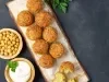 Fotografia vista de cima de oito unidades de falafel sobre um apoio de madeira claro retangular, ao lado de um falafel aberto também sobre o apoio. Ao lado do apoio tem dois recipientes de madeira com grão de bico e um molho de iogurte.