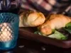 Fotografia em tons escuros duas empanadas recheadas com sardinha sobrepostas e apoiadas em uma tábua de madeira retangular escura. Na frente da tábua tem um copo de vidro com texturas de cor azul com uma vela dentro.