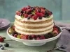Fotografia em tons de vermelho e lilás em uma bancada de madeira verde, um pano roxo, um suporte para bolo com o bolo pelado (naked cake) com creme branco e com decoração de frutas vermelhas. Ao fundo, pratos pequenos lilás de sobremesa.