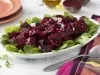 Fotografia em tons de roxo em uma bancada de madeira branca com um pano colorido xadrez com roxo, rosa e amarelo. Ao centro, um recipiente branco oval com salada de beterraba temperada com salsinha e cebola. Ao fundo, beterraba cortada ao meio.