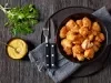 Fotografia vista de cima de um prato preto fundo sobre um pano dobrado cinza escuro, apoiado sobre um sousplat grande preto, ao lado de um ramo de salsinha e um pote com molho. Dentro do prato tem pedaços de frango empanado, ao lado tem talheres.