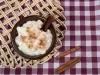 Fotografia vista de cima de um recipiente pequeno de vidro marrom com uma receita de canjica vegana. O recipiente está sobre um pano de mesa quadriculado de tom vinho e branco, ao lado do recipiente tem três paus de canela.