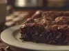 Fotografia de um pedaço de brownie de chocolate em um prato pequeno, branco de vidro raso. O prato está sobre uma mesa de madeira. Ao lado do prato tem uma xícara de tom claro.