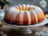 Fotografia de um bolo bundt dourado, polvilhado com açúcar de confeiteiro, em um suporte decorativo. O fundo sugere um ambiente acolhedor, com bananas e flores.