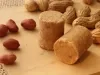 Fotografia em tons de caramelo com uma paçoca de formato cilíndrico ao centro. Em volta da paçoca existe alguns amendoins