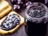 Fotografia de um recipiente de vidro com geleia de jabuticaba sobre uma mesa de madeira escura. Ao lado do recipiente tem algumas torradas, e uma delas está com um pouco de geleia, ao lado também de algumas jabuticabas.