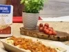 Fotografia em tons de marrom com uma travessa funda ao centro. Dentro da travessa existe algumas panquecas recheadas de carne cobertas com molho de tomate. Ao fundo existe um ramo de tomate cereja e uma embalagem de Aveia Flocos NESTLÉ.