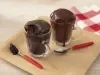 Foto da receita de bolo de chocolate de caneca feito com nescau extra cacau servida em duas canecas de vidro acompanhadas de duas colheres de cabo laranja sobre uma tábua de madeira. Esses elementos sobre uma mesa de madeira com um pano quadriculado