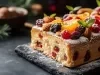 Fotografia de uma fatia de bolo retangular de Natal decorado com frutas secas, castanhas e folhas de hortelã, polvilhado com açúcar de confeiteiro e apresentado em uma base de pedra escura.
