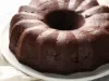 Bolo de chocolate bundt, com cor marrom escuro, repousa em um prato branco. A superfície brilhante e as ranhuras verticais criam um padrão atraente.