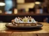 Em cima de uma mesa, prato com fatia de banoffee com massa crocante, doce de leite com chantilly, decorado com chantilly, bananas em rodelas e raspas de chocolate.