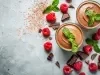 Fotografia vista de cima de dois potes de vidro sobre uma mesa de cimento queimado. Dentro do copos tem a sobremesa, finalizada com ganache e folhas de hortelã. Ao redor dos copos tem hortelãs, pedaços de chocolate e framboesas espalhadas.