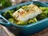 Em cima de uma mesa, refratário azul escuro com posta de bacalhau e floretes de brócolis.