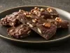 Em cima de uma mesa, barra com pedaços de massa com nozes, chocolate derretido e nozes picadas.