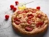 Fotografia de uma focaccia com tomate cereja assado, sal grosso e azeite. Ao lado da focaccia tem um recipiente de vidro pequeno com azeite, ao lado alguns tomates cereja, sobre uma mesa branca com texturas.