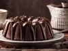 Em cima de uma mesa, prato redondo com bolo de chocolate com furo no meio e cobertura de chocolate cremoso.