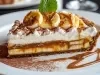 Em cima de uma mesa, prato com fatia de banoffee com massa crocante, doce de leite cremoso, decorado com chantilly, bananas em rodelas e raspas de chocolate e uma folha de hortelã.