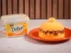 Foto de um cuscuz recheado com charque, servido em um prato laranja, ao lado de uma embalagem de margarina deline