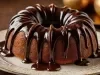 Fotografia de bolo de bundt dourado, apresentado em um prato decorativo. Coberto com um lustroso glacê de chocolate, o bolo é emoldurado por um fundo festivo com esferas douradas.