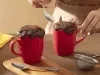 Fotografia de duas canecas vermelhas com bolo e cobertura de nescau extra cacau, ao lado de uma colher de sopa e uma mini peneira. A foto mostra uma mão femnina segurando uma das canecas.