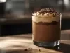 Fotografia da sobremesa com o creme com doce de leite e chocolate, kitkat e raspas de chocolate por cima, dentro de um copo de vidro pequeno, sobre uma mesa de madeira.