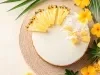 Fotografia de torta de abacaxi vista de cima em tons claros de uma torta inteira feita com massa de bolo. A torta estpa decorada com abacaxis em pedaços e cubos, com coco ralado. Ao redor tem algumas flores amarelas e plantas.