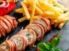 Fotografia em tons claros de dois tornedores de frango recheados enrolados no bacon, ao redor tem tomates, cebola roxa fatiada, batatas fritas e algumas folhas, sobre um apoio quadrado preto. O apoio está sobre uma mesa de madeira.