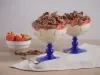 Foto da receita de pavê de leite moça com morangos e chocolate, servidos em taças com uma base azul, sobre uma bancada de madeira decorada com um tecido, pedaços de chocolate, um pote com morangos e uma lata de Leite Moça