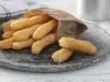 Imagem de um prato rústico escuro com um saquinho de papel marrom com palitos de biscoito de polvilho dourados.