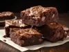 Fotografia de alguns pedaços de brownie com gotas de chocolate sobrepostos, apoiados em uma folha branca, apoiada em uma mesa de madeira.