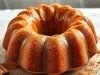 Fotografia de um bolo bundt dourado, colocado em um prato branco. O bolo apresenta uma textura úmida e é acompanhado por canela e uma xícara de bebida quente.