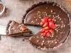 Fotografia vista de cima de uma torta grande com base de chocolate, recheio de creme branco e ganache por cima, decorada com morangos e castanhas trituradas. A foto mostra uma faca grande segurando uma fatia da torta.